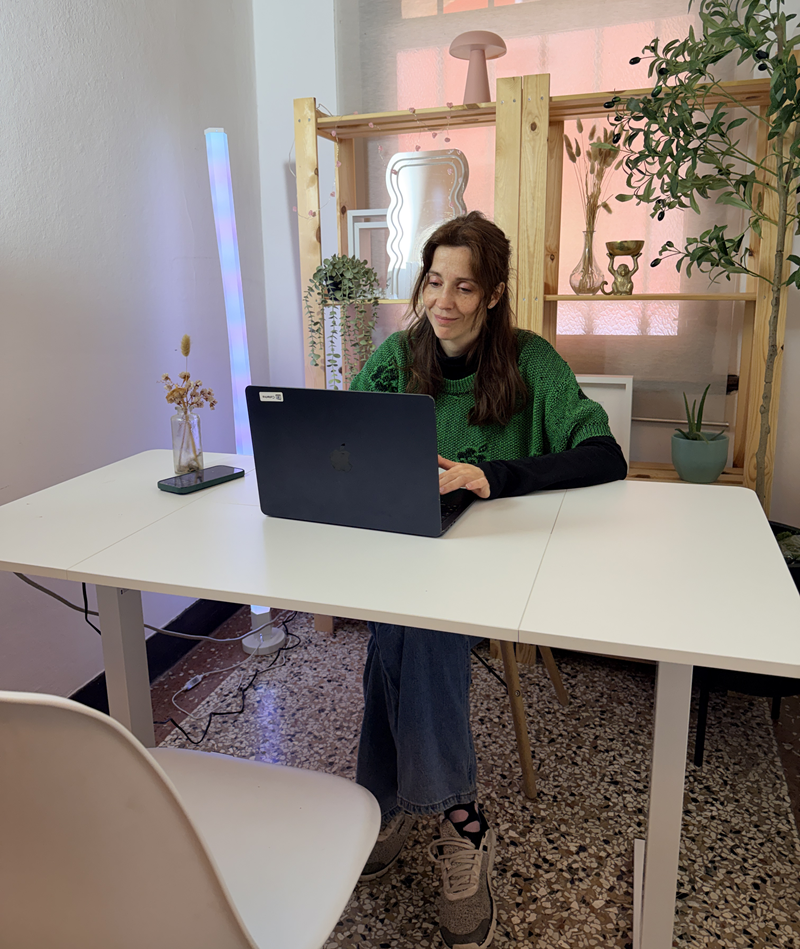 Donna con maglione verde al laptop su scrivania bianca, scaffale in legno con piante e pavimento alla veneziana