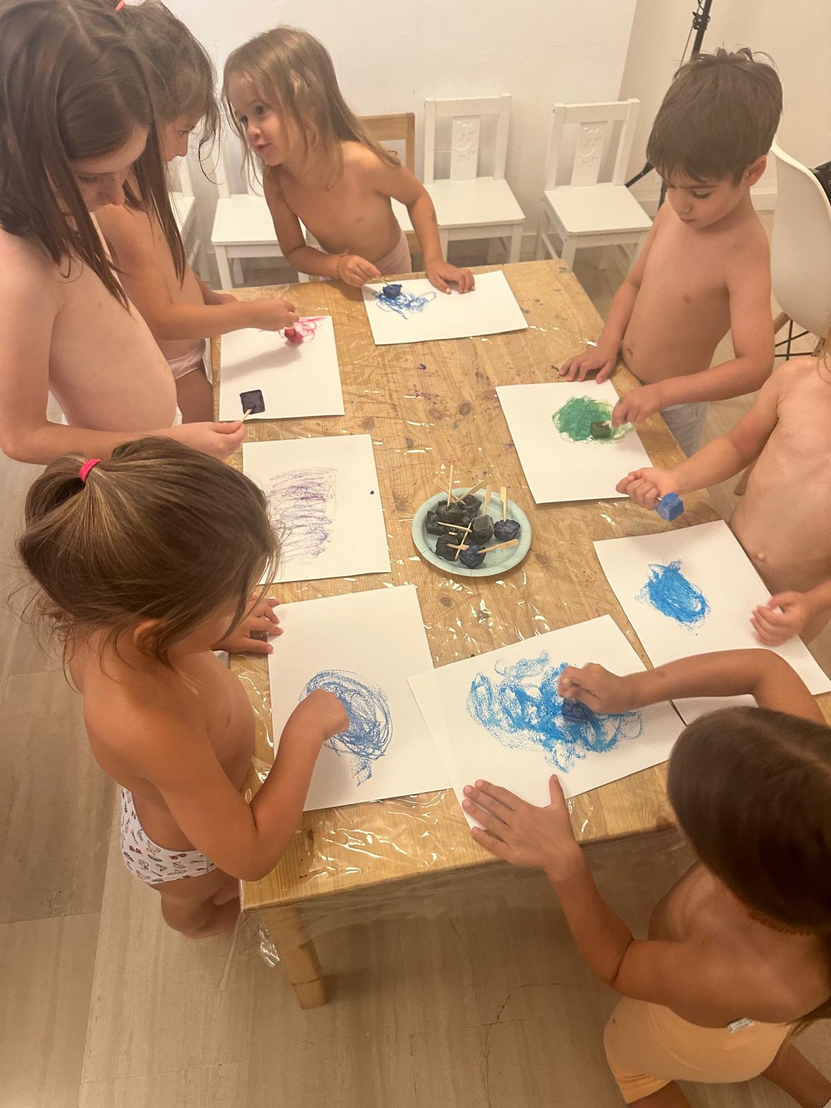 Bambini attorno a un tavolo di legno coperto da cellophane colorano fogli bianchi con colori a ghiacciolo blu, verdi e viola
