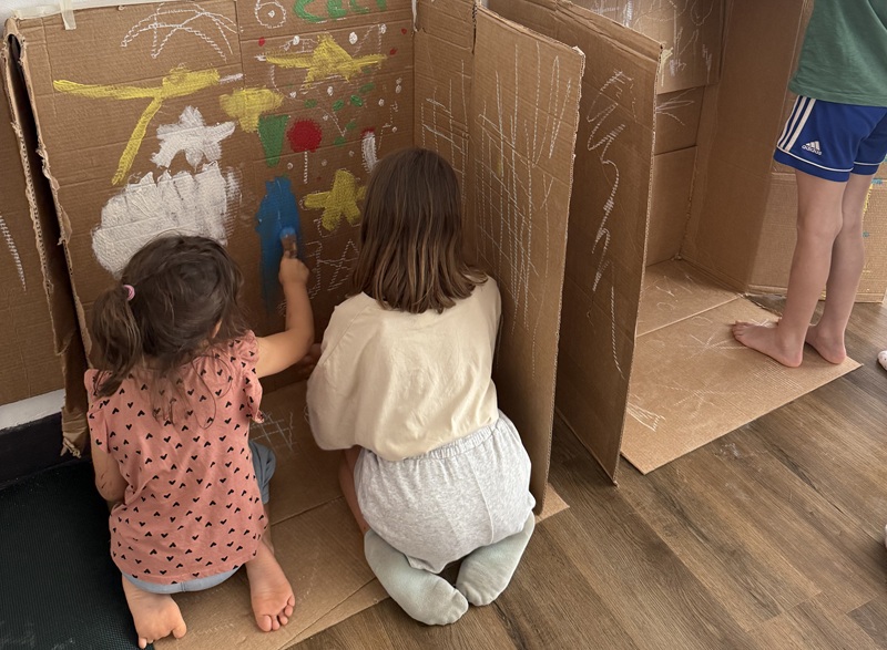 Due bambine in ginocchio dipingono con gessi e pennello pannelli di cartone, su pavimento in legno, accanto a un bimbo scalzo