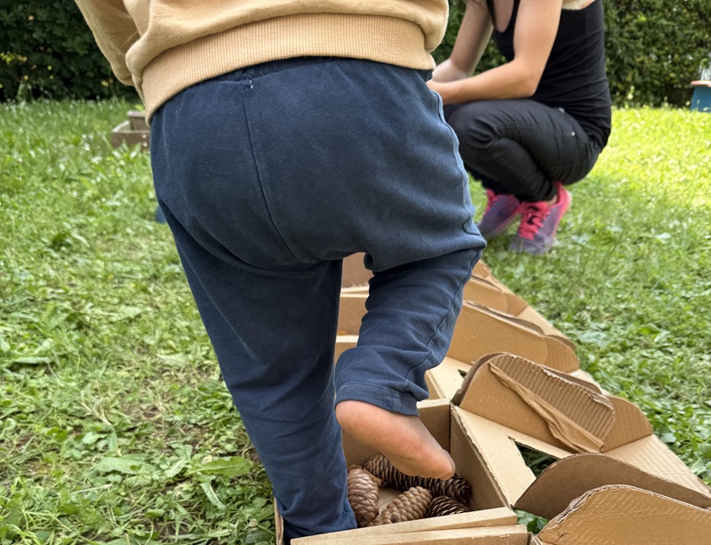 Bambino scalzo in pantaloni blu cammina su un percorso sensoriale di scatole di cartone con pigne, sul prato del Yoyo Summer
